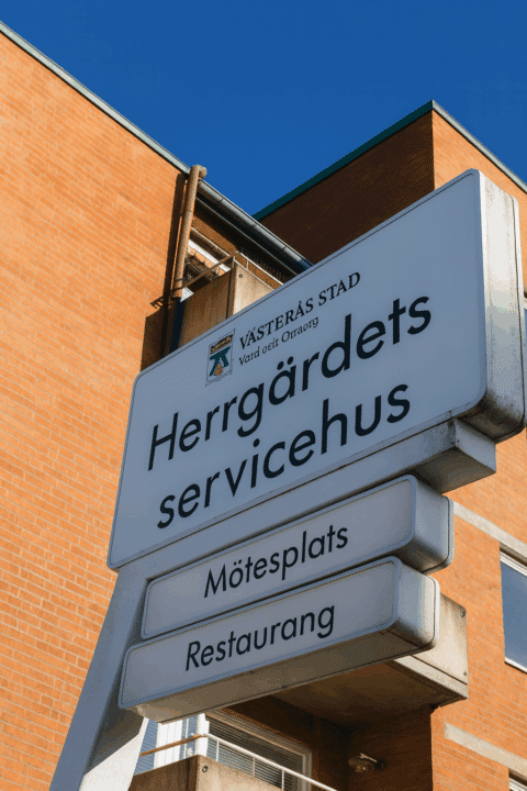 Vägskylt för Herrgärdets servicehus i Västerås, med text om mötesplats och restaurang, mot bakgrund av en röd tegelbyggnad.
